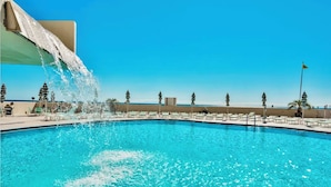 A swimming pool with a water feature, surrounded by lounge chairs and umbrellas, with a clear view of the ocean in the background.