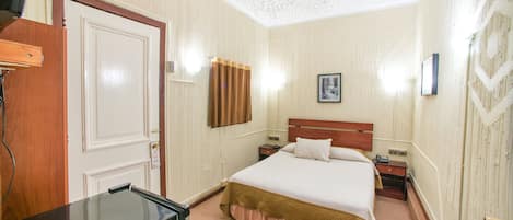 A hotel room with a wooden door, a bed with a white and brown bedspread, a nightstand, and a wall-mounted light.