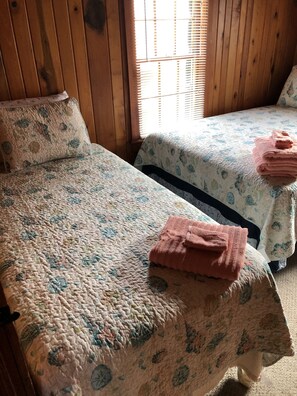 Two single beds with floral bedding and pink towels, wooden walls, and a window with blinds. - Calico Jack's Galley (Kitty Hawk)