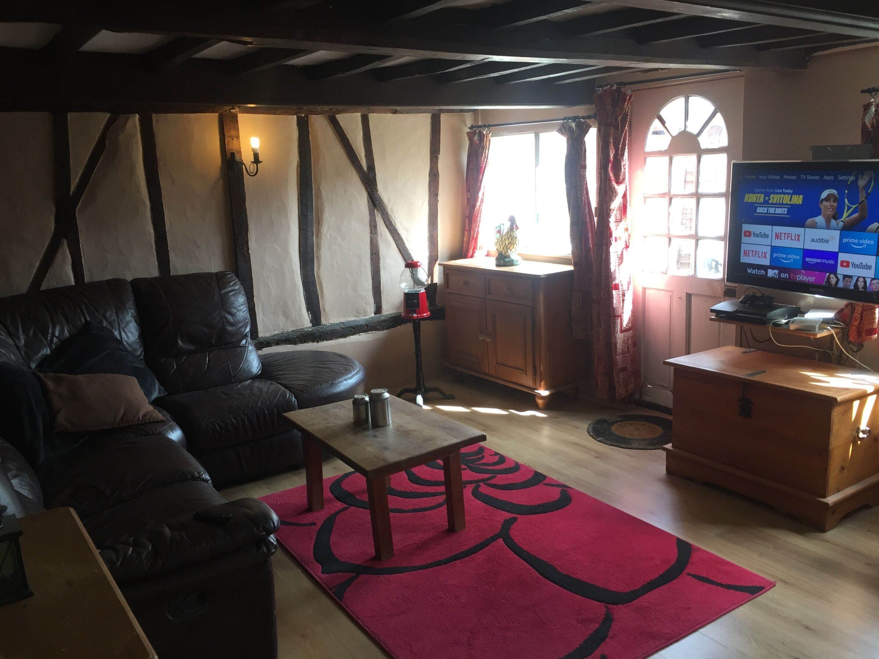 A cozy living room with a leather sofa, a wooden coffee table, and a red rug. There's a TV on a wooden stand, a window with curtains, and a wooden cabinet.