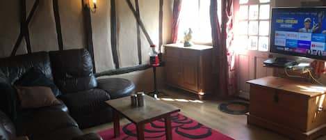 A cozy living room with a leather sofa, a wooden coffee table, and a red rug. There's a TV on a wooden stand, a window with curtains, and a wooden cabinet.