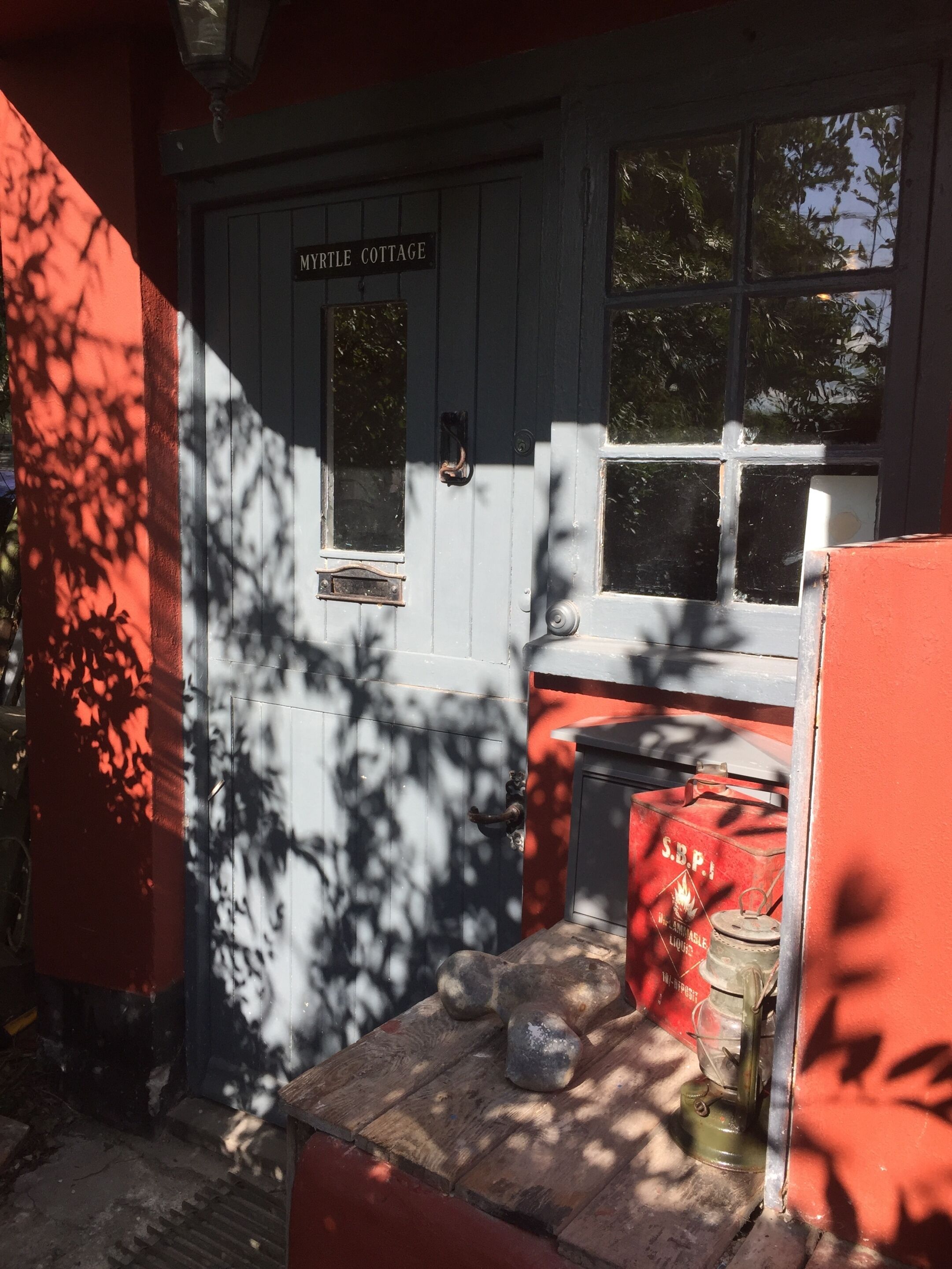 A cottage entrance with a sign reading "Myrtle Cottage," a wooden door, and a small table with a red object on it.