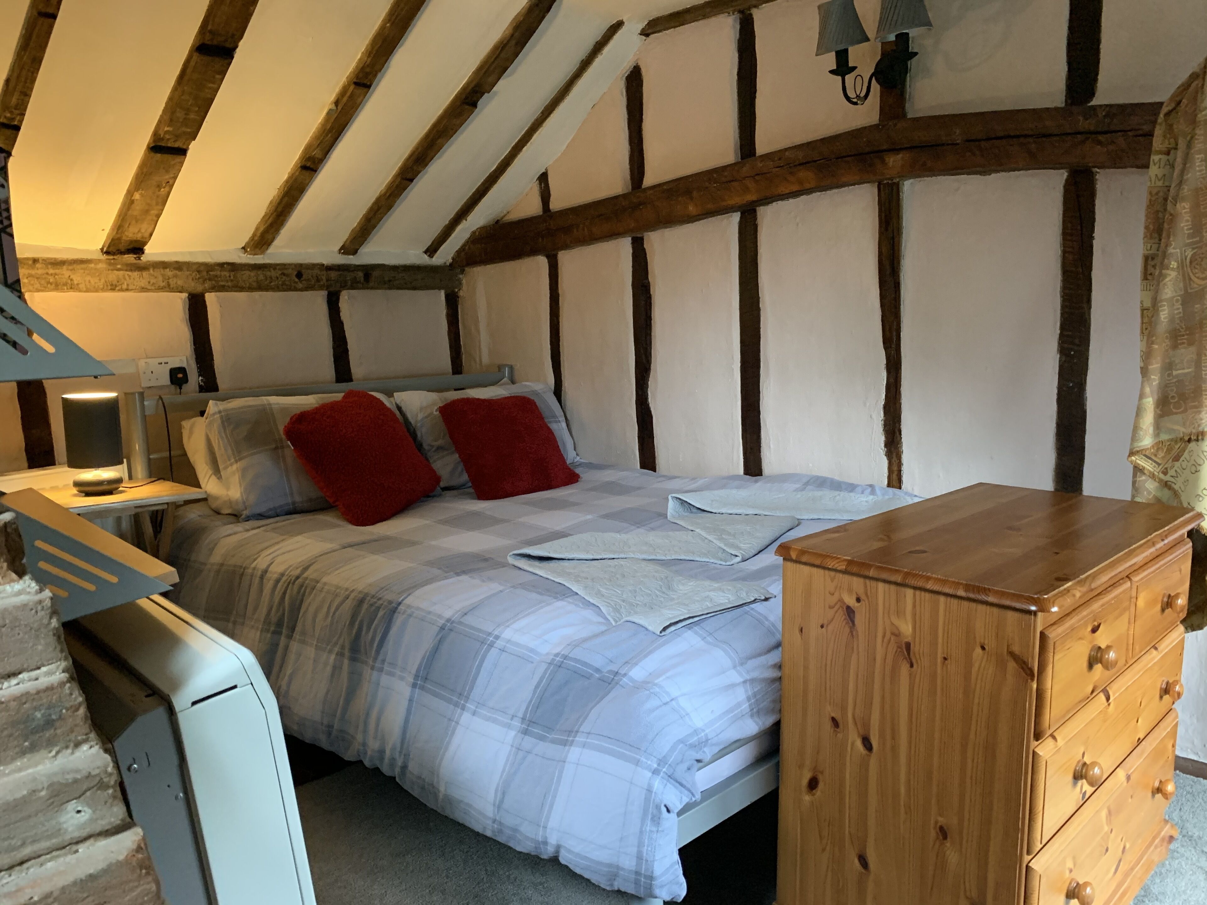 Een slaapkamer met een bed, een houten dressoir, een bureau met lamp en een schuine plafond met zichtbare balken.