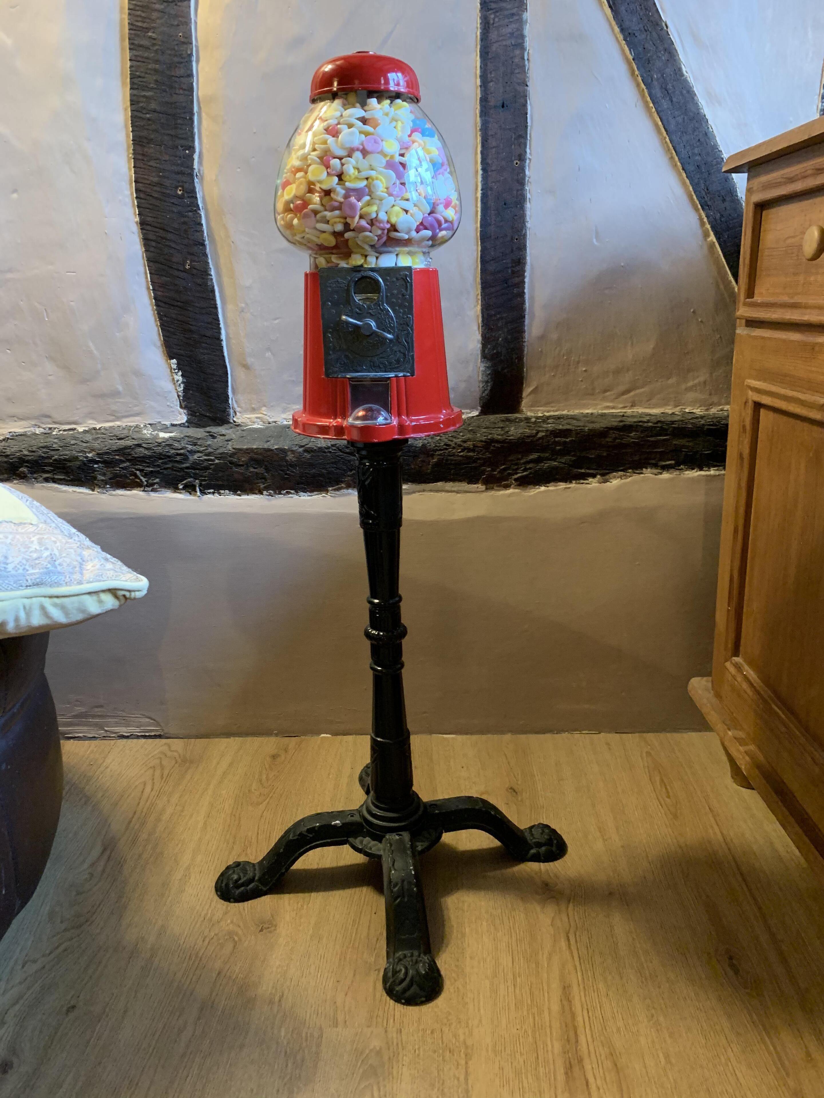 Um distribuidor de doces antigo sobre um suporte de metal preto com topo vermelho, colocado sobre um piso de madeira em frente a uma parede com vigas aparentes.