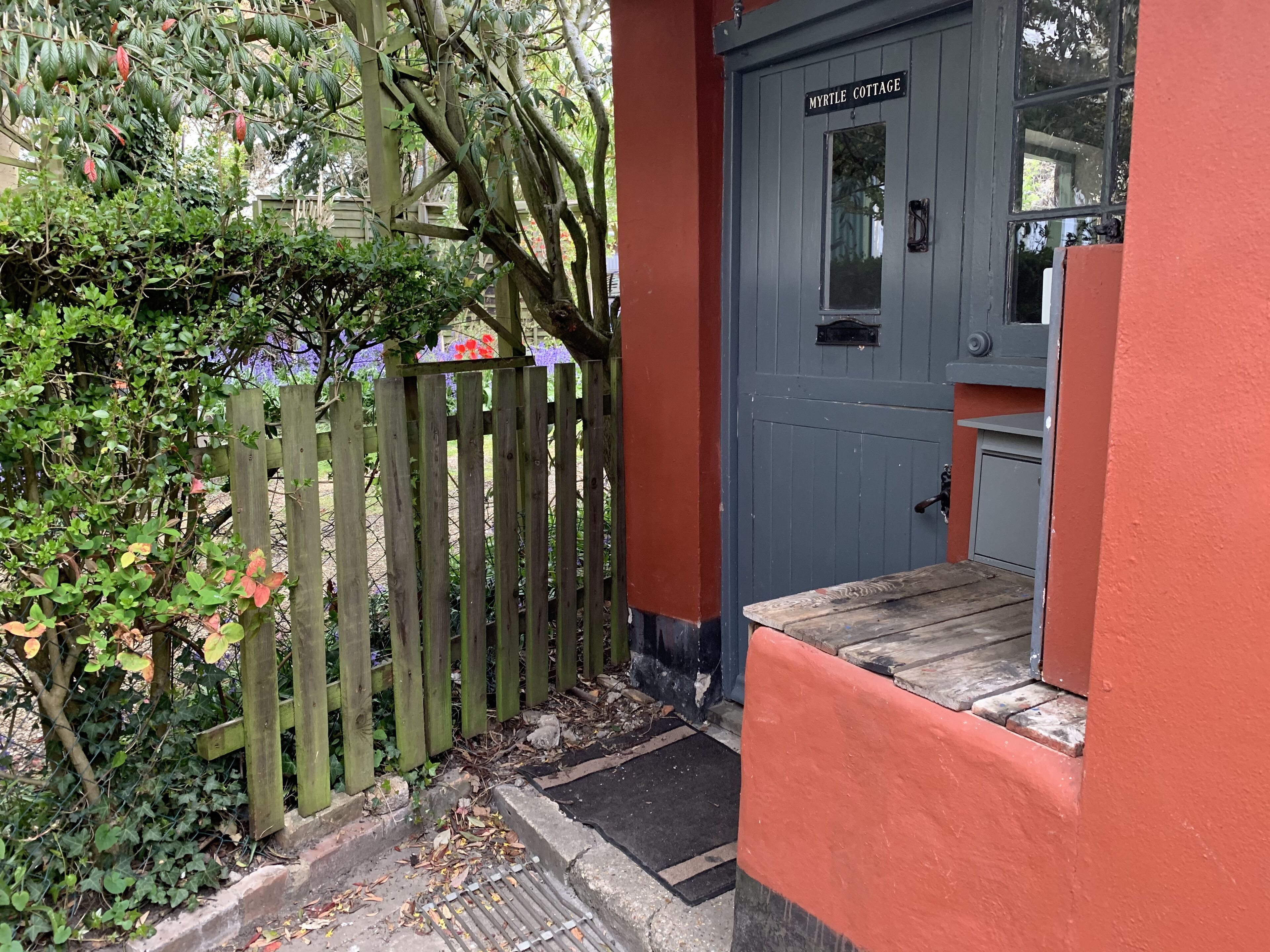 Una verja de madera, una puerta gris con la leyenda "MYRTLE COTTAGE" y una pared roja.