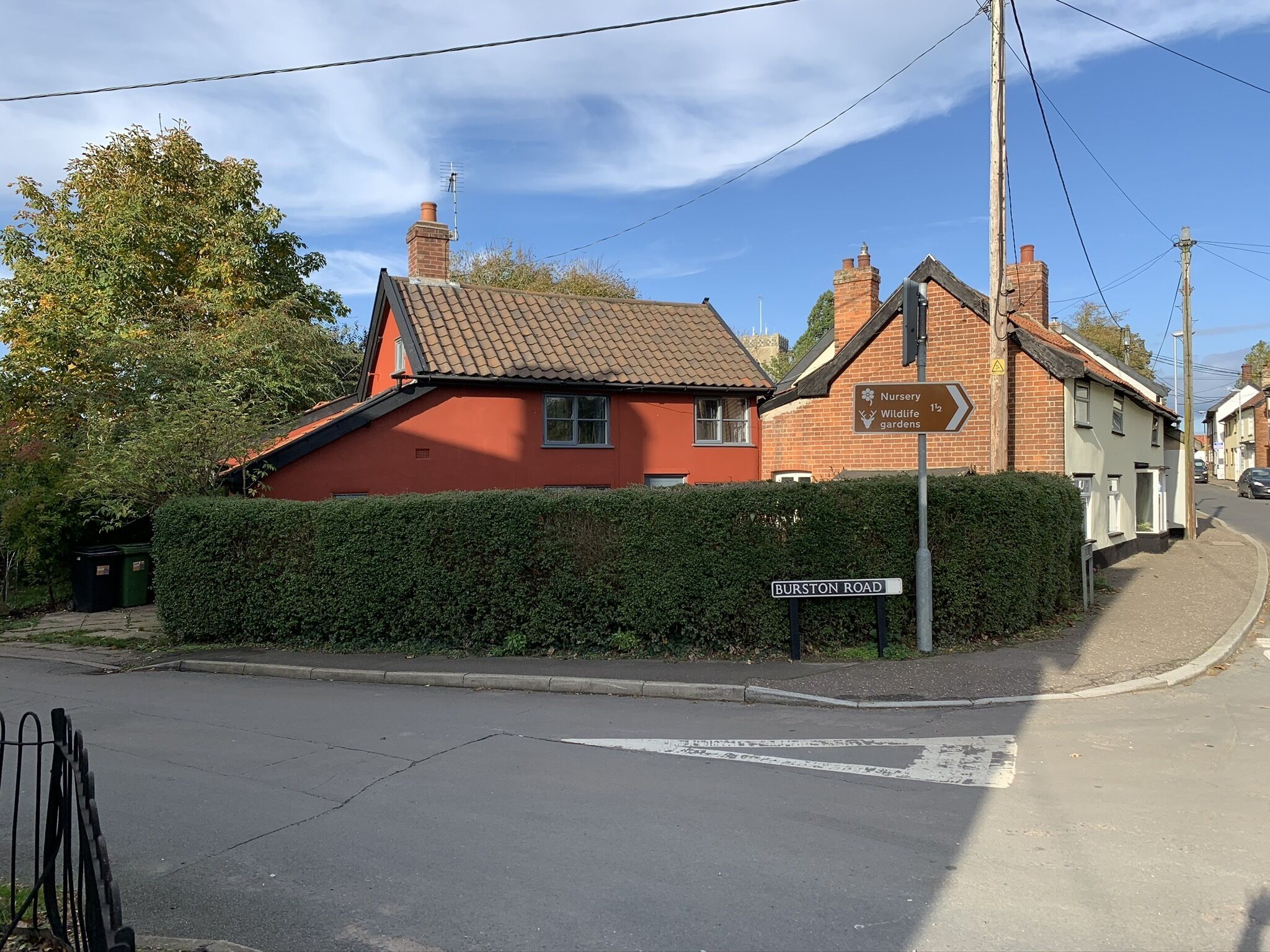 A residential street with houses, a sign for a nursery, wildlife gardens, and a street name sign for Burston Road.