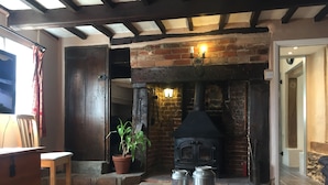A rustic room with a brick fireplace, wooden beams, a potted plant, and metal containers on the floor. - Myrtle Cottage (Diss)
