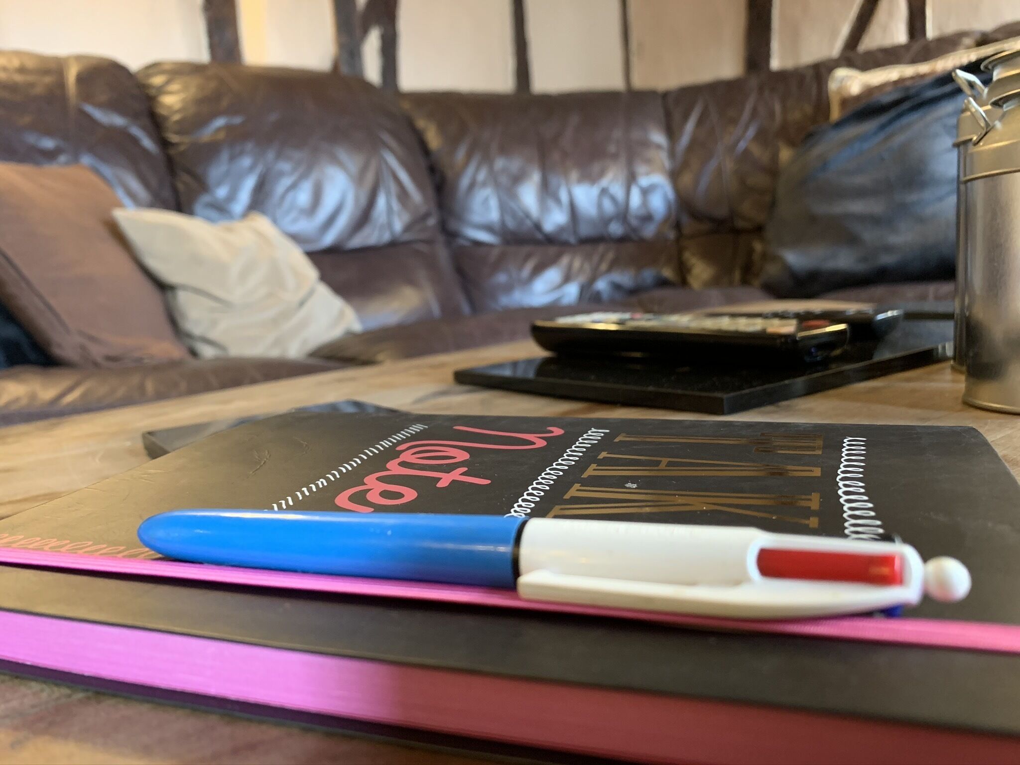 A notebook with a blue pen on a pink mat, a black object, and a metal container on a wooden table.