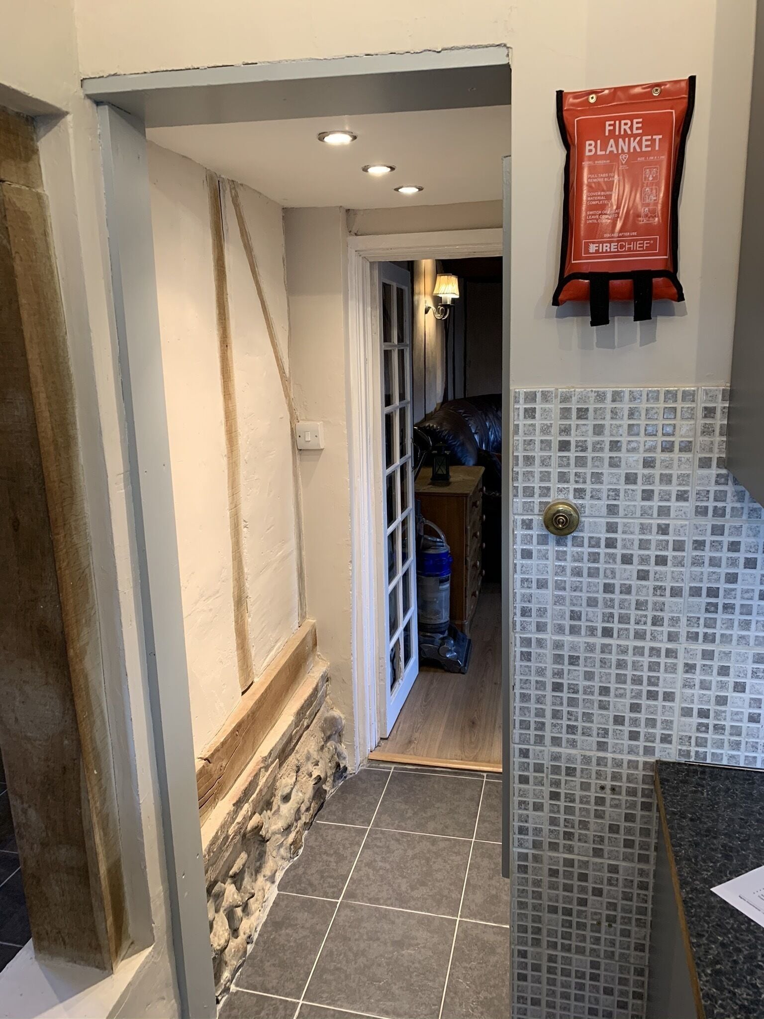 Un couloir étroit au sol carrelé, une couverture anti-feu accrochée au mur et une porte donnant sur une pièce avec un lit et un mur également carrelé.
