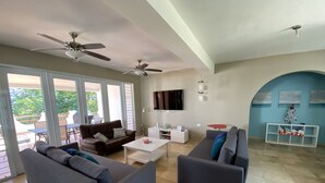 Uma sala de estar espaçosa com um sofá cinza, uma mesa de centro branca e uma poltrona cinza. Há uma TV de tela plana na parede, um ventilador de teto e um pequeno aparador branco com objetos decorativos.