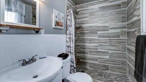 A bathroom with a white sink, toilet, and shower area with grey tiles. - Cedar Village Motel (Spokane)