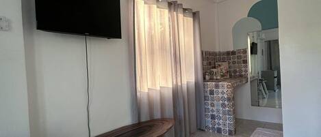 Una sala de estar con un televisor de pantalla plana montado en la pared, una mesa de madera con un control remoto y un juego de cortinas.