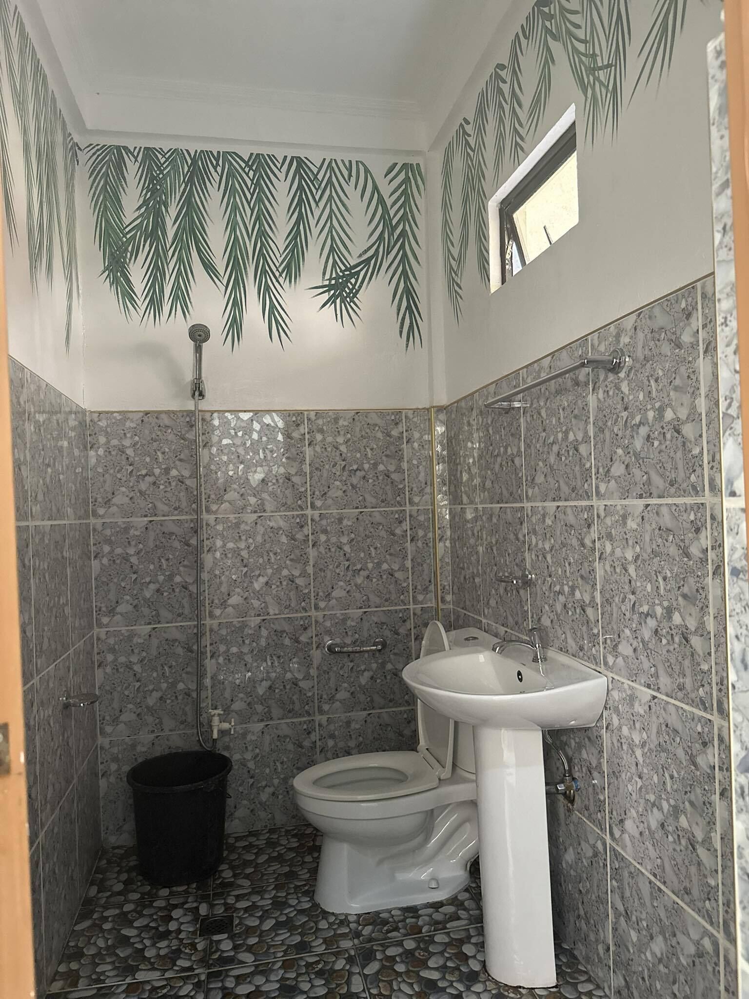 Une salle de bain équipée d’une douche, de toilettes et d’un lavabo, avec un sol recouvert de petits galets et un mur orné d’un motif de feuilles.