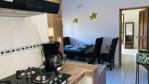 Et køkken med komfur, kaffemaskine og et spiseområde med stole og bord.