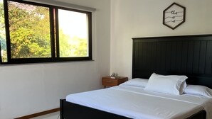 Une chambre avec un lit, une table de chevet, un ventilateur de plafond, une fenêtre donnant sur un paysage verdoyant et un élément décoratif fixé au mur.