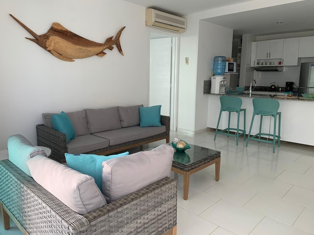 Una sala moderna con un sofá gris, cojines color turquesa, un sillón de mimbre y una mesa de centro. Se ve una cocina al fondo con gabinetes blancos y taburetes de bar color turquesa.