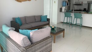 Un salon moderne avec un canapé gris, des coussins turquoise, un fauteuil en osier et une table basse. On aperçoit, au fond, une cuisine avec des meubles blancs et des tabourets de bar turquoise.