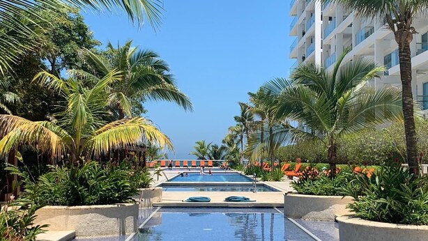 Une piscine entourée de chaises longues et de palmiers, le tout bordé d’un immeuble doté de balcons.