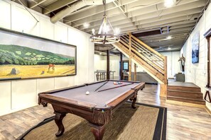 Une table de billard dans une pièce ornée d’un grand tableau, d’un escalier en bois et d’un lustre.