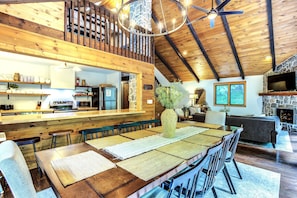 Un vaste espace dédié à la salle à manger, avec une longue table en bois, de nombreuses chaises, une cheminée en pierre et un plafond haut avec des poutres apparentes.