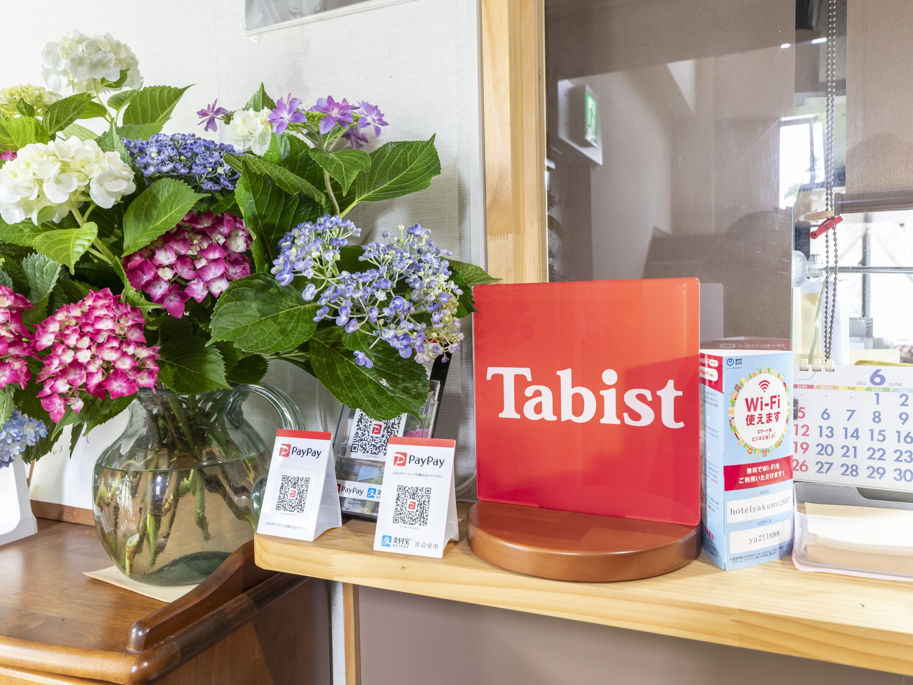 A hotel reception desk with a vase of flowers, a red 'Tabist' sign, a PayPay payment sign, and a calendar.