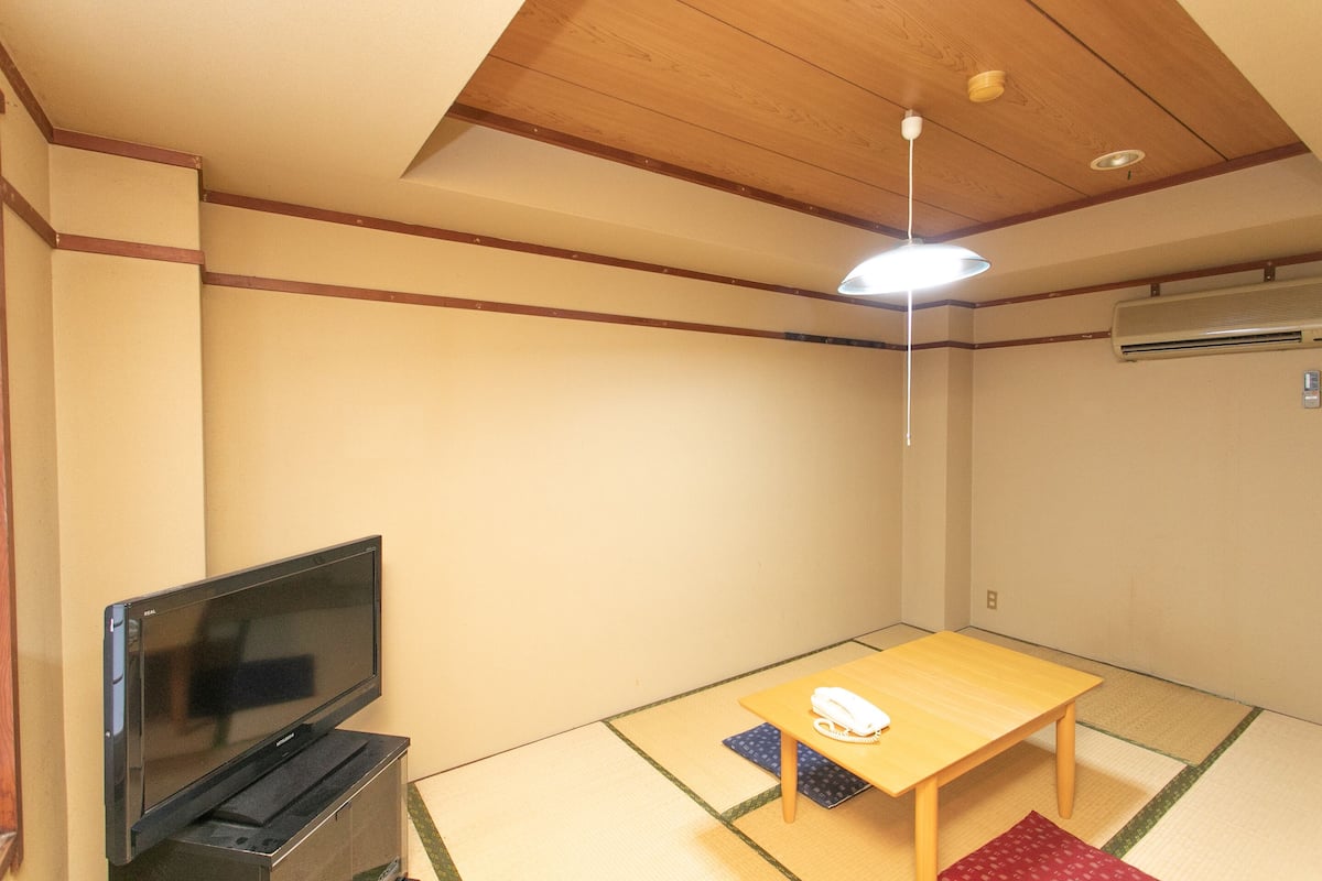 Ein Zimmer mit Tatami-Boden, einem Fernseher, einem Tisch mit Telefon und einer Deckenleuchte.