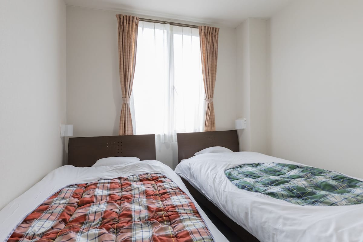 Ein Zimmer mit zwei Einzelbetten, jedes mit einer verschiedenen Bettdecke. Es gibt ein Fenster mit Vorhängen, eine Wandlampe und eine Steckdose.