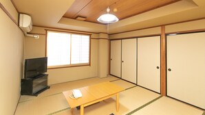Une pièce de style japonais traditionnel, avec un sol recouvert de tatamis, une basse table en bois, un téléviseur posé sur un meuble, des portes coulissantes et une fenêtre à motifs géométriques.