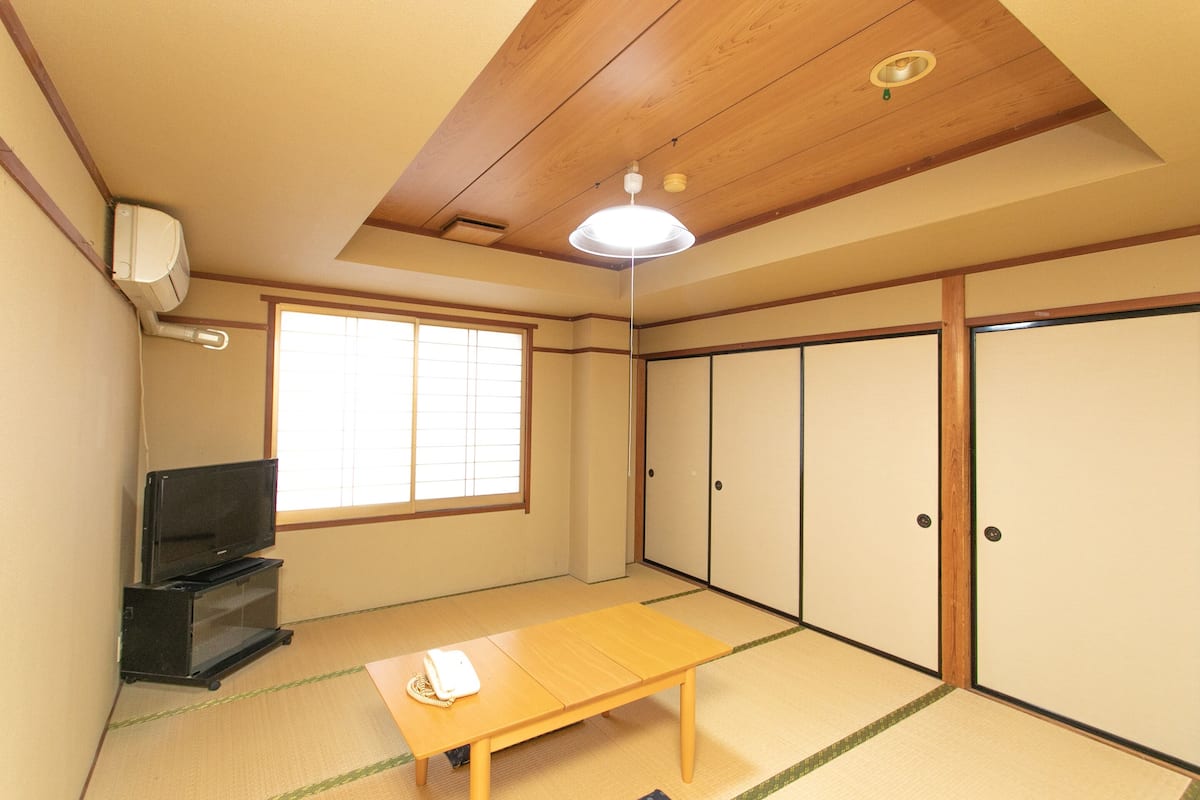 Ein traditioneller japanischer Raum mit Tatami-Bodenbelag, einem niedrigen Holztisch, einem Fernsehgerät auf einem Standfuß, Schiebetüren und einem Fenster mit Gittermuster.
