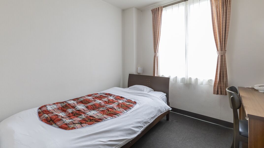 A single bed with a plaid blanket, a bedside table, a chair, and a window with curtains.