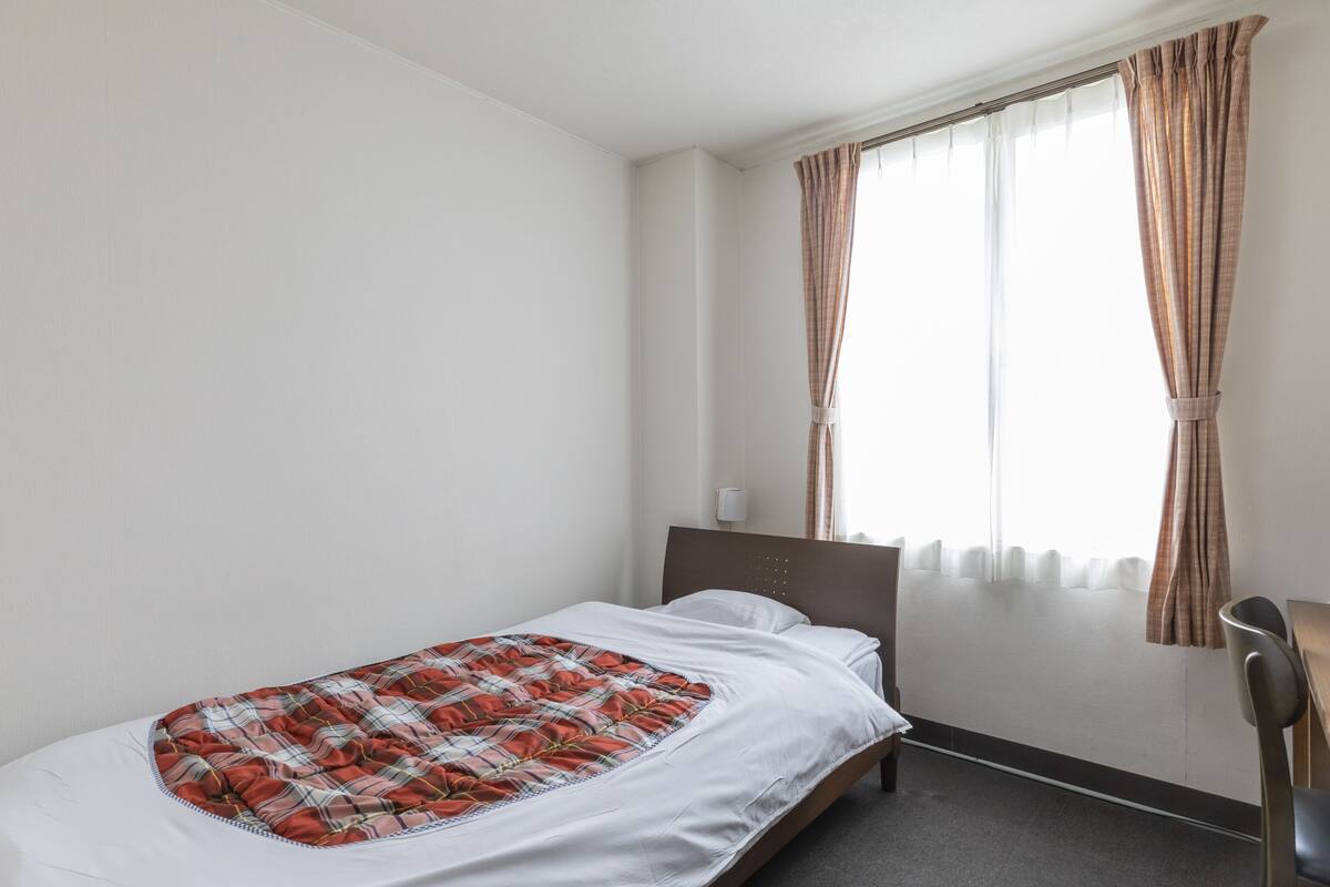 Ein Einzelbett mit kariertem Überwurf, ein Nachttisch, ein Stuhl und ein Fenster mit Vorhängen.