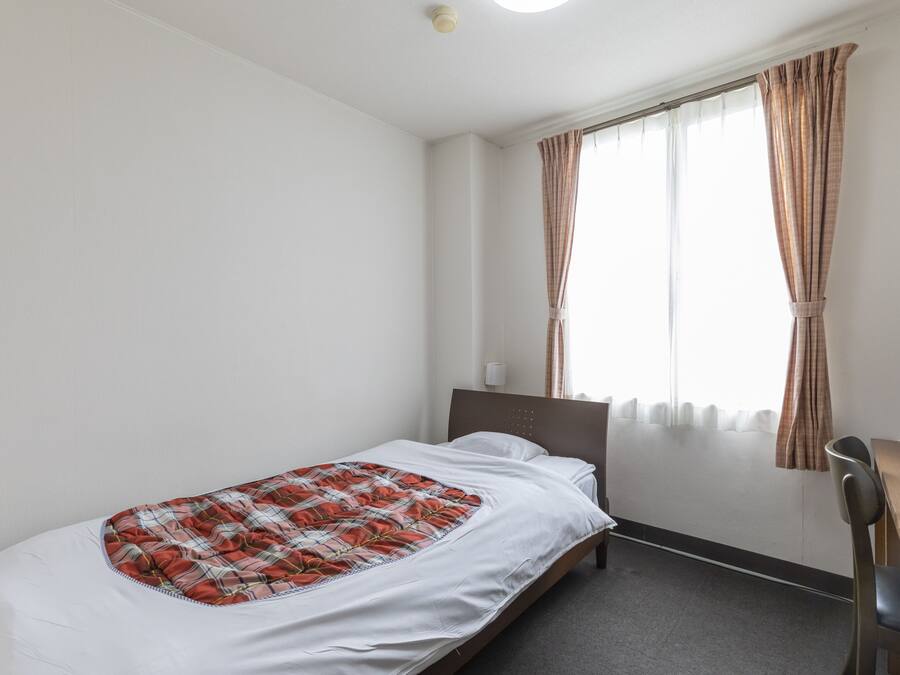 A single bed with a plaid blanket, a bedside table, a chair, and a window with curtains.