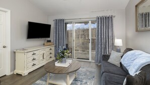 Un salon équipé d’un téléviseur à écran plat, d’une commode blanche, d’un canapé gris et d’une porte-fenêtre coulissante donnant sur un espace extérieur.