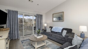 Un salon avec un canapé, une table basse et un téléviseur. Il y a un balcon avec des rideaux et une vue sur l’extérieur.