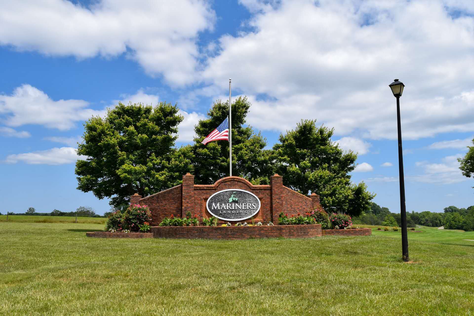 A brick sign with 'MARINERS LANDING' and an American flag, surrounded by greenery and a clear sky.