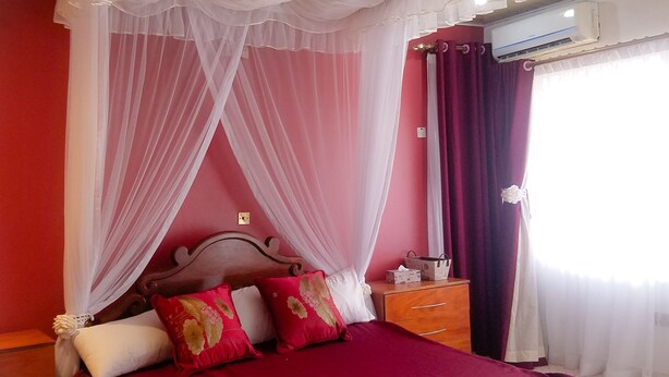 Un lit avec une couvre-lit rouge, une tête de lit en bois et un baldaquin en tissu blanc transparent.