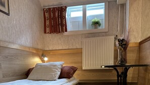 A compact room with a bed, a small table, a window with curtains, a radiator, and a plant. - The Red Boat (Stockholm)