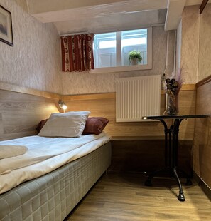 A compact room with a bed, a small table, a window with curtains, a radiator, and a plant. - The Red Boat (Stockholm)