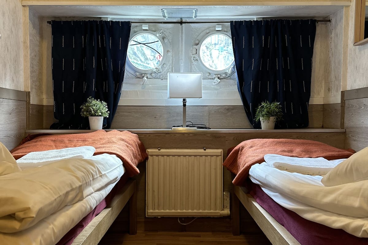 A room with two beds, a radiator, a window with curtains, a lamp, and a small plant.