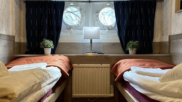 A room with two beds, a radiator, a window with curtains, a lamp, and a small plant. - The Red Boat (Stockholm)