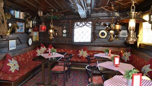 A rustic interior with wooden beams, red upholstered seating, and vintage decor. Tables are set with checkered tablecloths and potted plants. - The Red Boat (Stockholm)