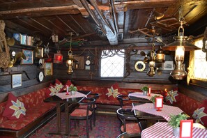 A rustic interior with wooden beams, red upholstered seating, and vintage decor. Tables are set with checkered tablecloths and potted plants. - The Red Boat (Stockholm)