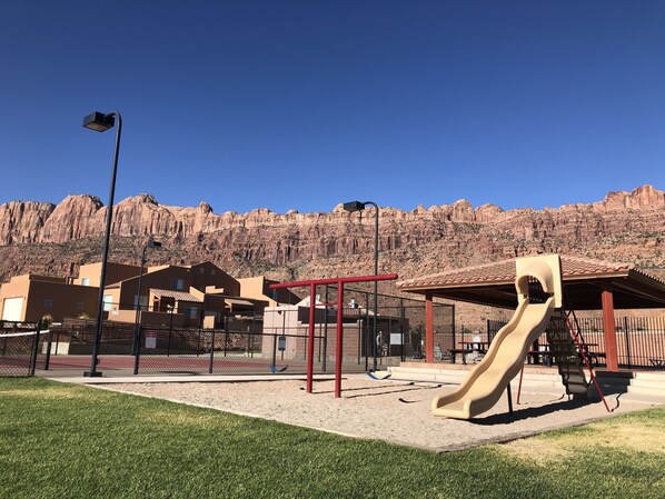 A playground with a slide and climbing structures, surrounded by buildings and a mountainous backdrop. - Moab Behind the Rocks Retreat - 3B/2.5B- Garage/Pool (Moab)