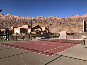 An outdoor tennis court with a red surface, surrounded by a fence, and a mountain backdrop.