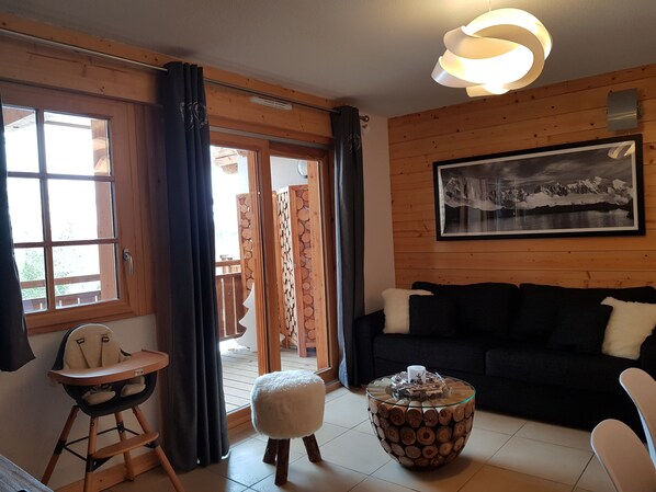 Un salon chaleureux avec un plancher en bois, un canapé noir, une table basse en verre, un petit tabouret moelleux blanc, une chaise haute et une grande fenêtre avec des rideaux.