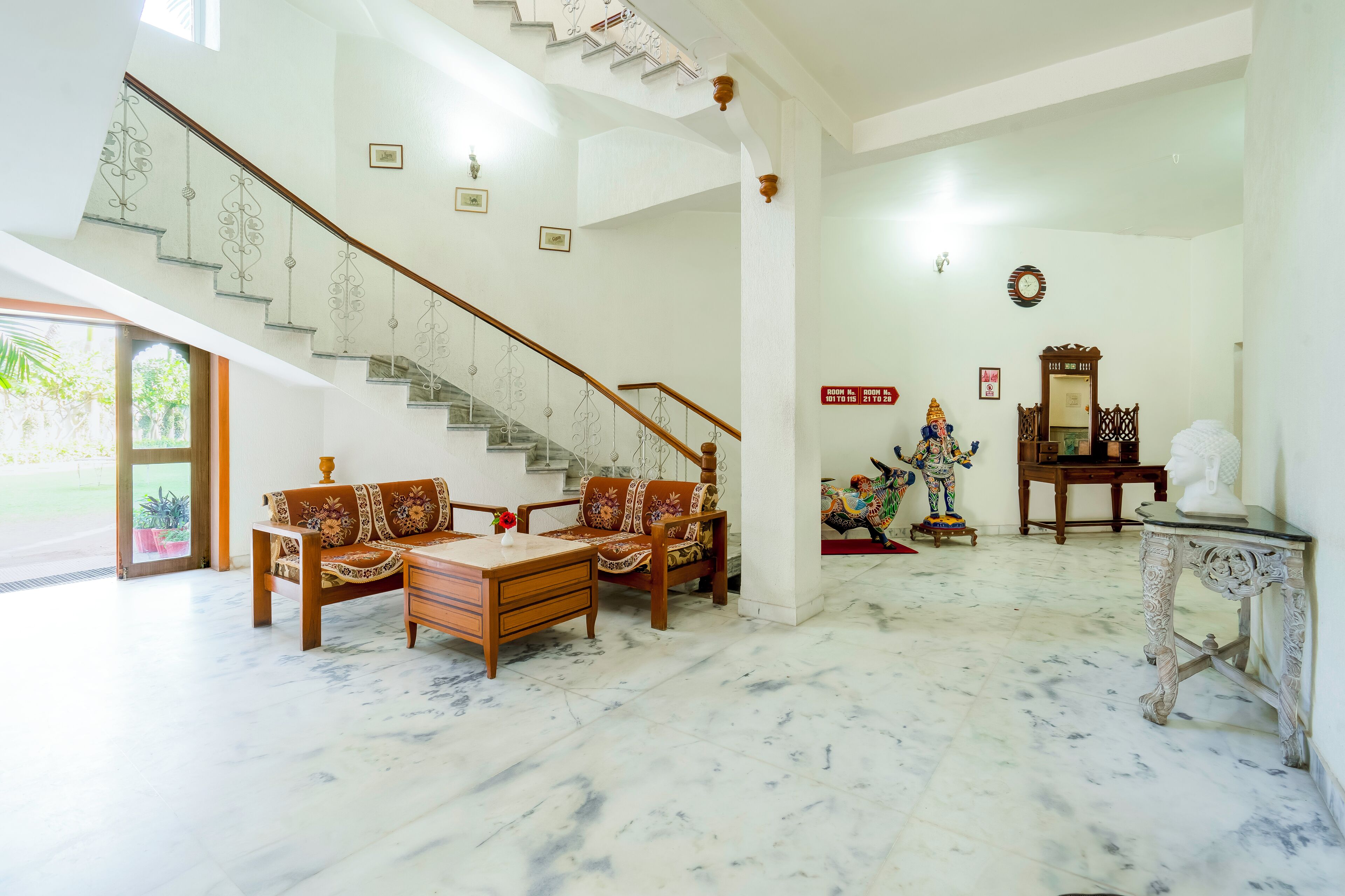A spacious living area with a marble floor, a wooden sofa set, a staircase with decorative railings, and a statue on the wall.
