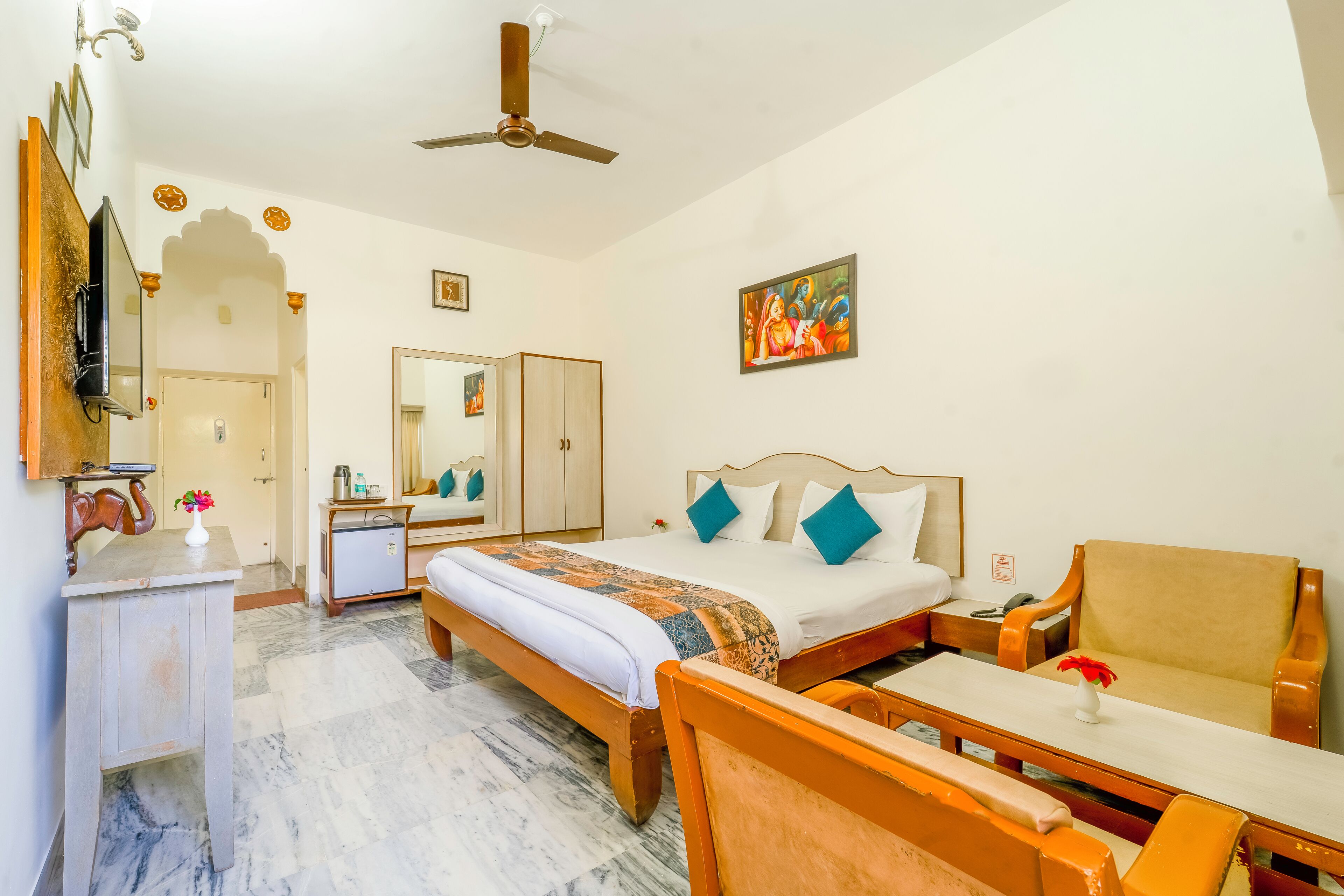 A hotel room with a bed, a desk, a chair, a ceiling fan, and a painting on the wall.