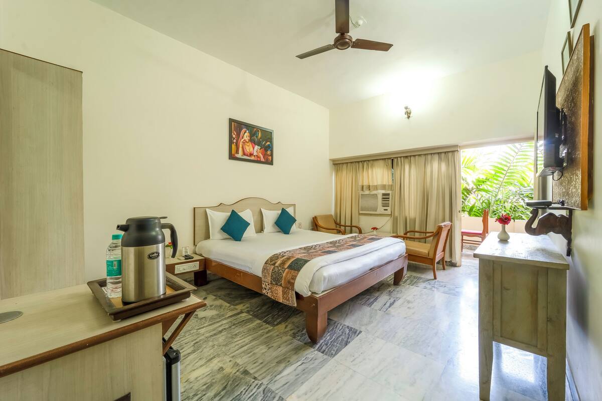 Une chambre d’hôtel avec un lit, un bureau, une chaise, un ventilateur de plafond, un tableau et une fenêtre avec des rideaux.