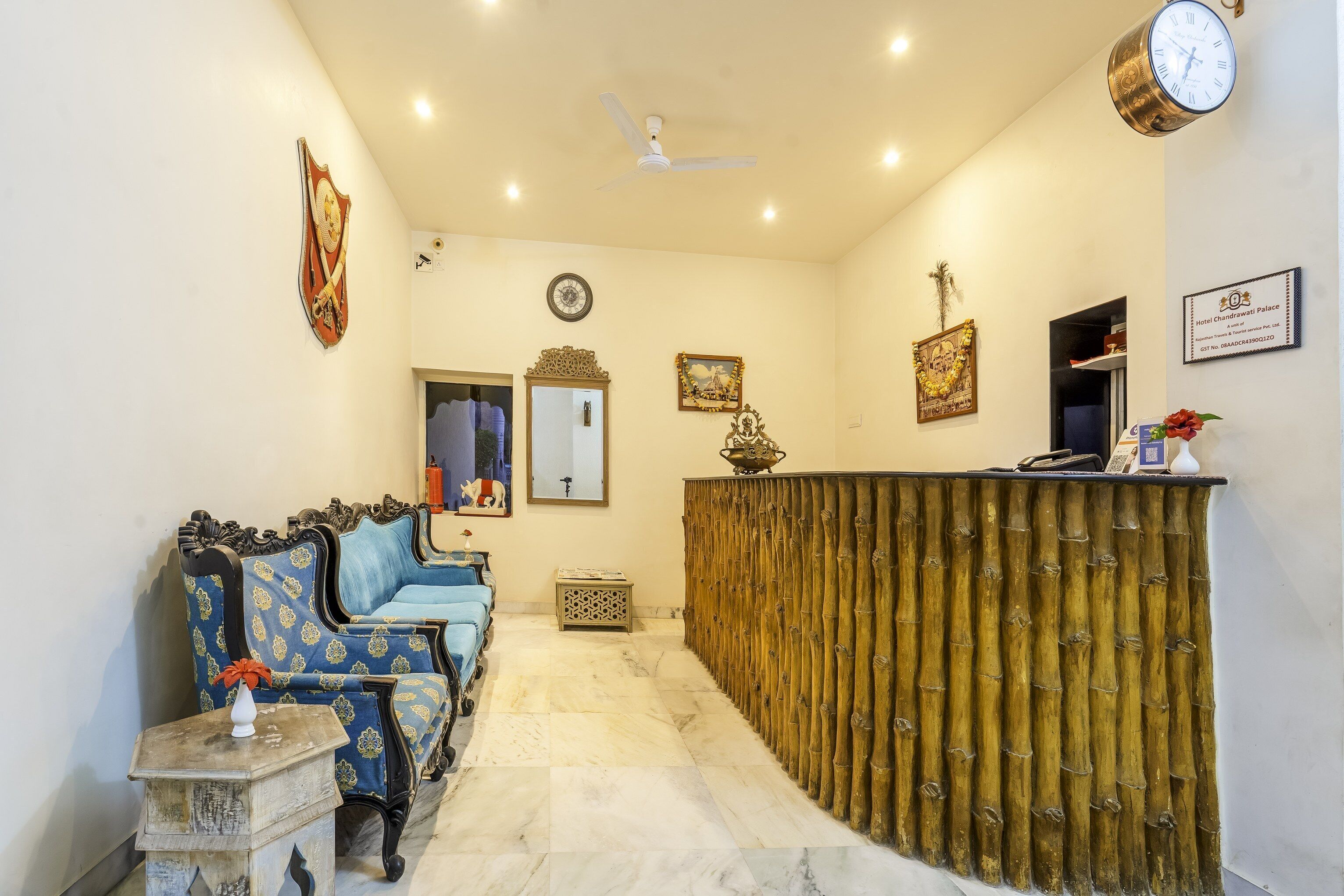 Un espace d’accueil comprenant un comptoir de réception, un canapé bleu à motifs, une petite table avec un vase et une horloge murale.