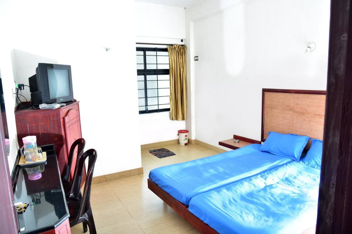 A bedroom with a bed, a desk with a television, a chair, a window with curtains, and a small table with items.